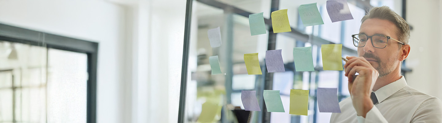 A man in an office, looking at colorful sticky notes on a glass wall and planning ideas.
