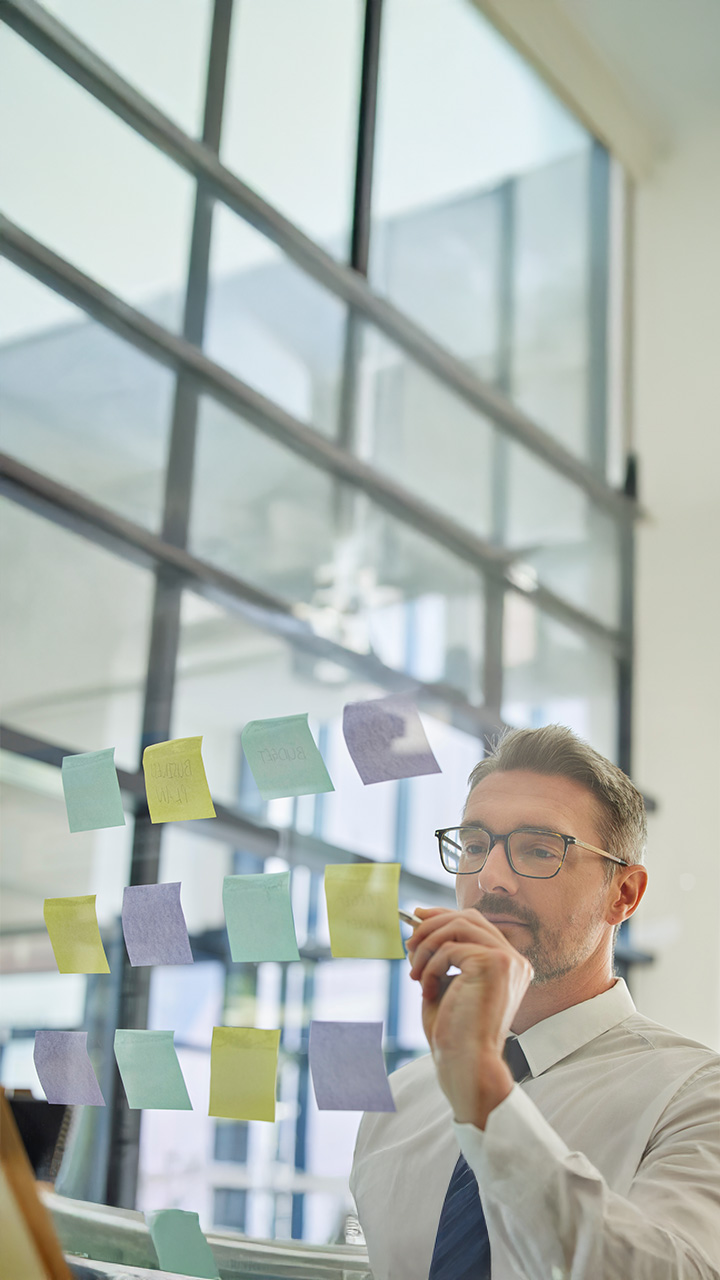 A man in an office, looking at colorful sticky notes on a glass wall and planning ideas.